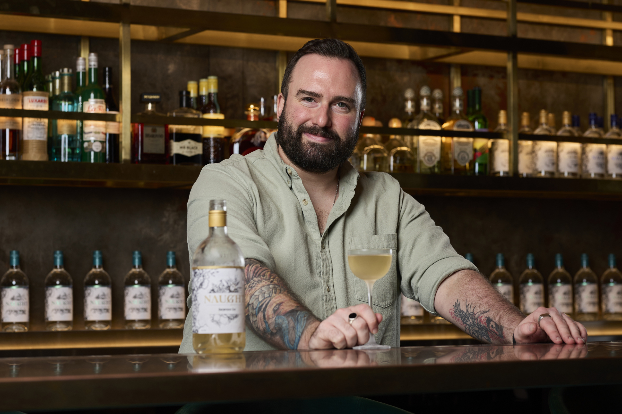 Man with tattoos and a beard sitting at a bar, smiling while holding a cocktail glass next to a bottle of Naught gin, with a backdrop of illuminated shelves filled with various spirits, creating a warm and inviting atmosphere.