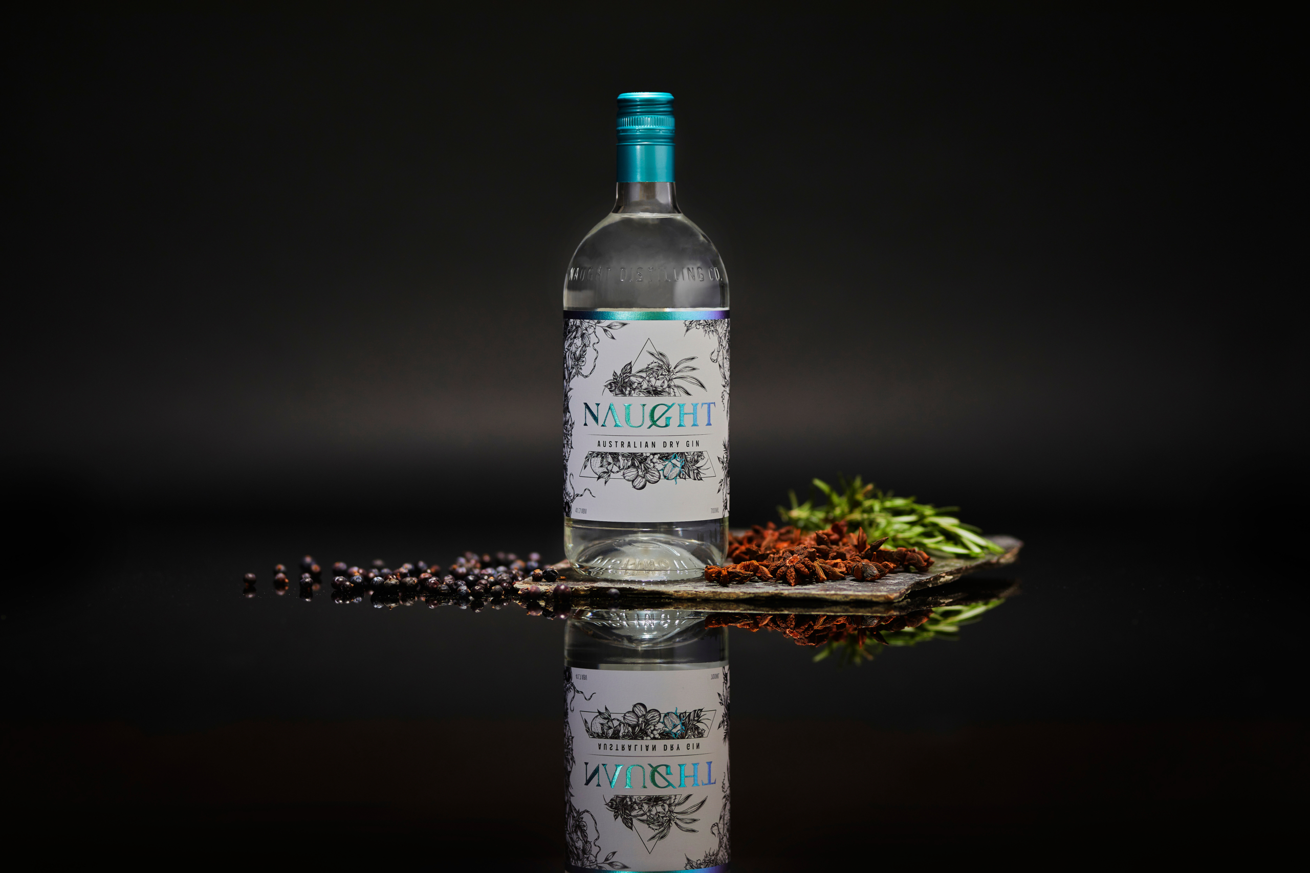 Australian Dry Gin bottle from Naught displayed with botanicals including juniper berries, star anise, and rosemary on a dark reflective surface, highlighting its artisanal design and premium quality.