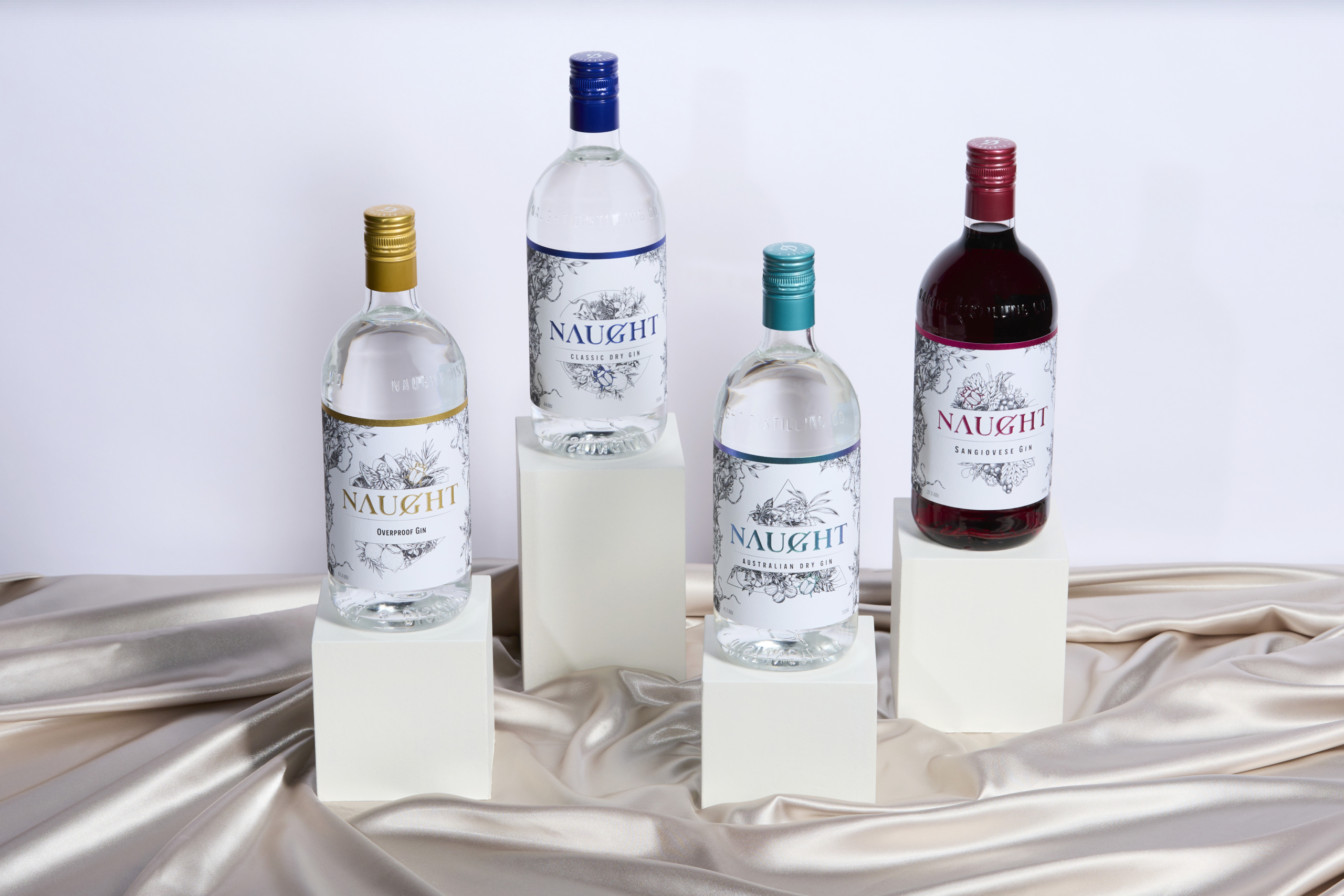 Four bottles of Naught gin varieties, including Overproof Gin, Classic Dry Gin, Australian Dry Gin, and Sangiovese Gin, elegantly displayed on white pedestals with a satin fabric background, showcasing the diversity and premium quality of Naught's gin selection