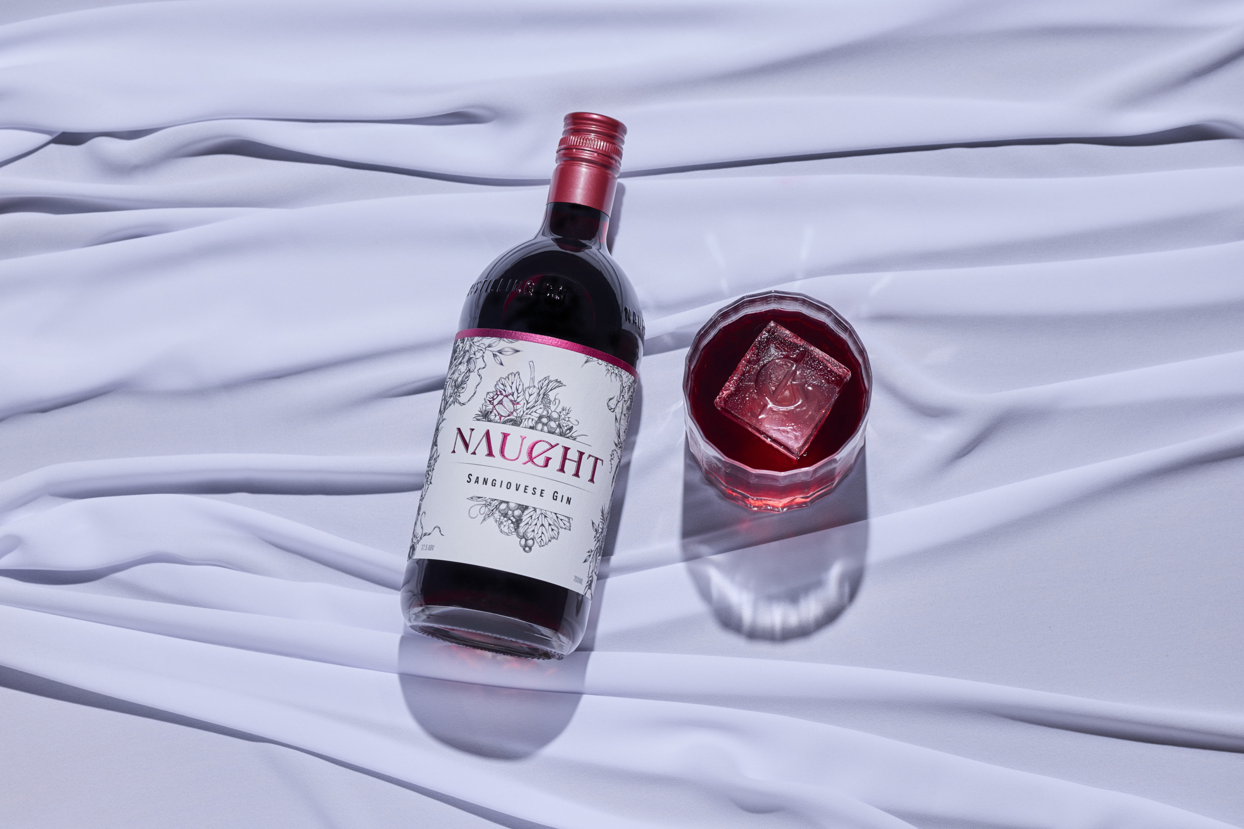 Naught Sangiovese Gin bottle laid on soft white fabric alongside a glass with a red gin cocktail and a large branded ice cube, showcasing the rich color and premium presentation of this unique gin variety.