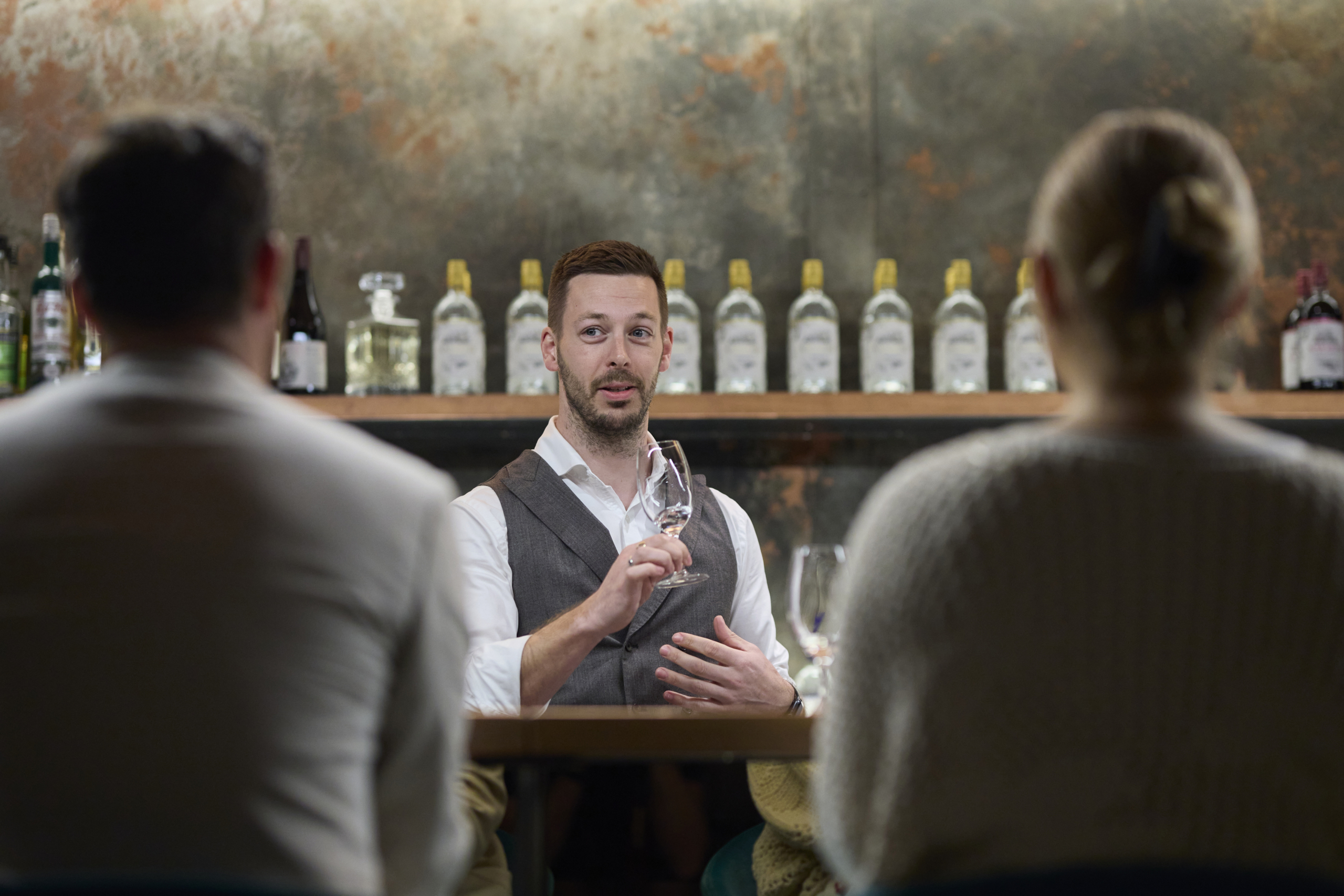 Man conducting a gin tasting session, holding a glass and speaking to attendees seated at a table, with a background of Naught gin bottles displayed on a shelf, emphasizing an engaging and educational experience