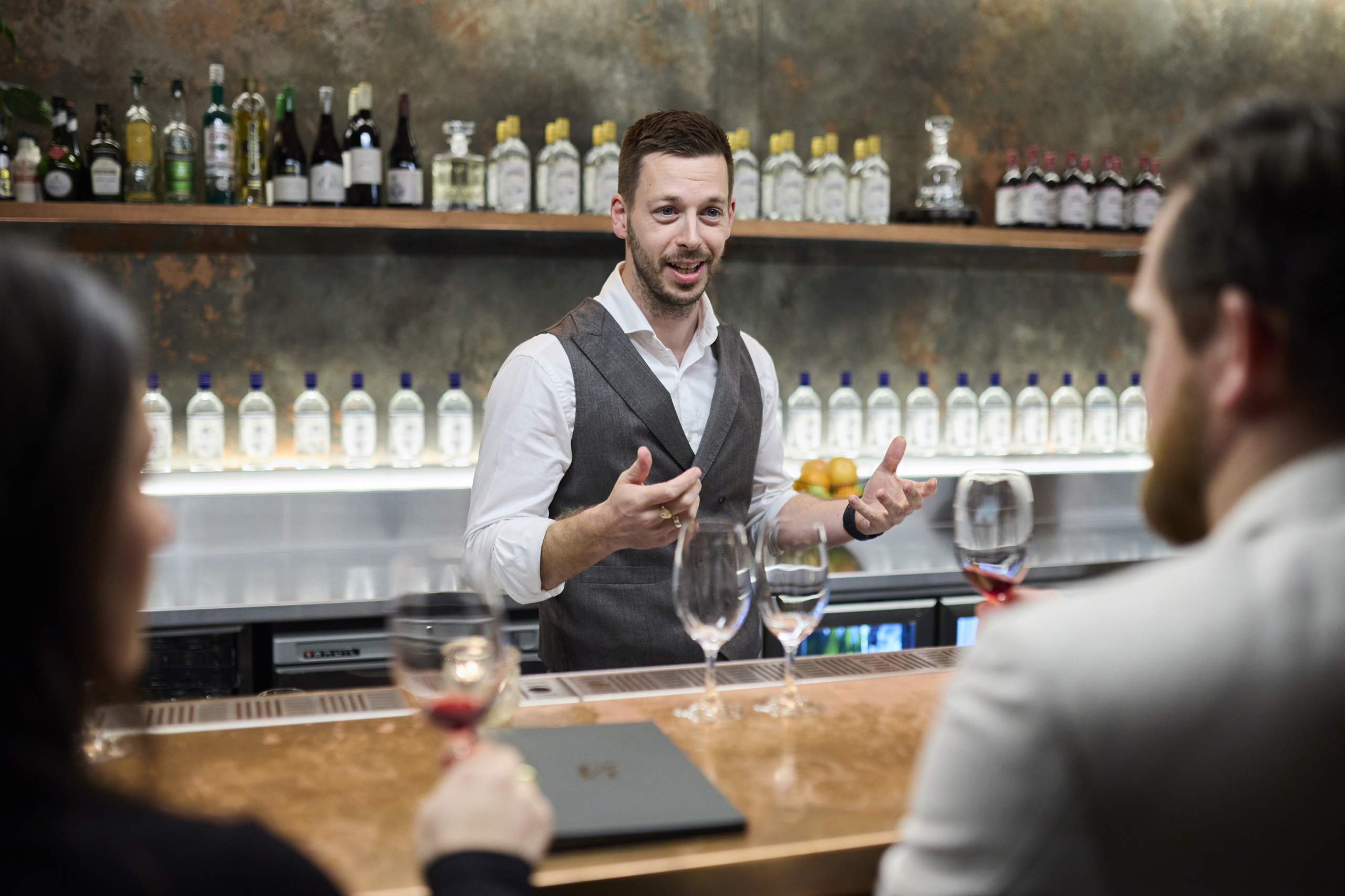 Man in a vest leading a gin tasting session behind a bar, engaging with two attendees holding wine glasses, with a backdrop of Naught gin bottles on shelves, creating an inviting and informative atmosphere.