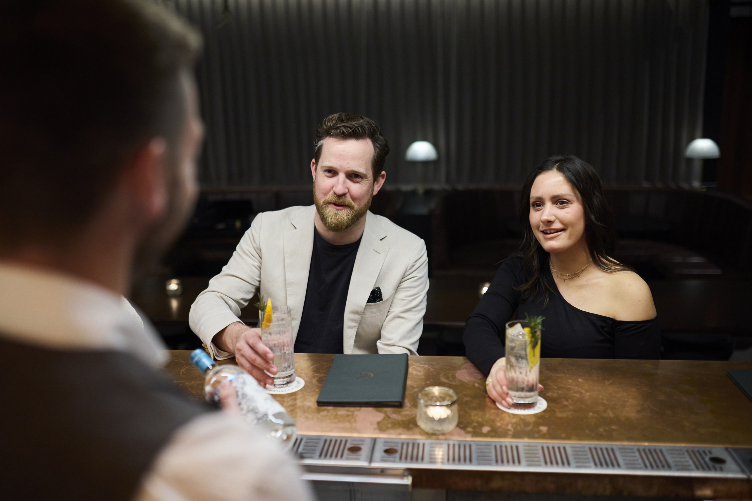 Two guests, a man in a beige blazer and a woman in a black top, seated at a bar enjoying cocktails, attentively listening to a bartender during a gin tasting session, creating a relaxed and engaging atmosphere.