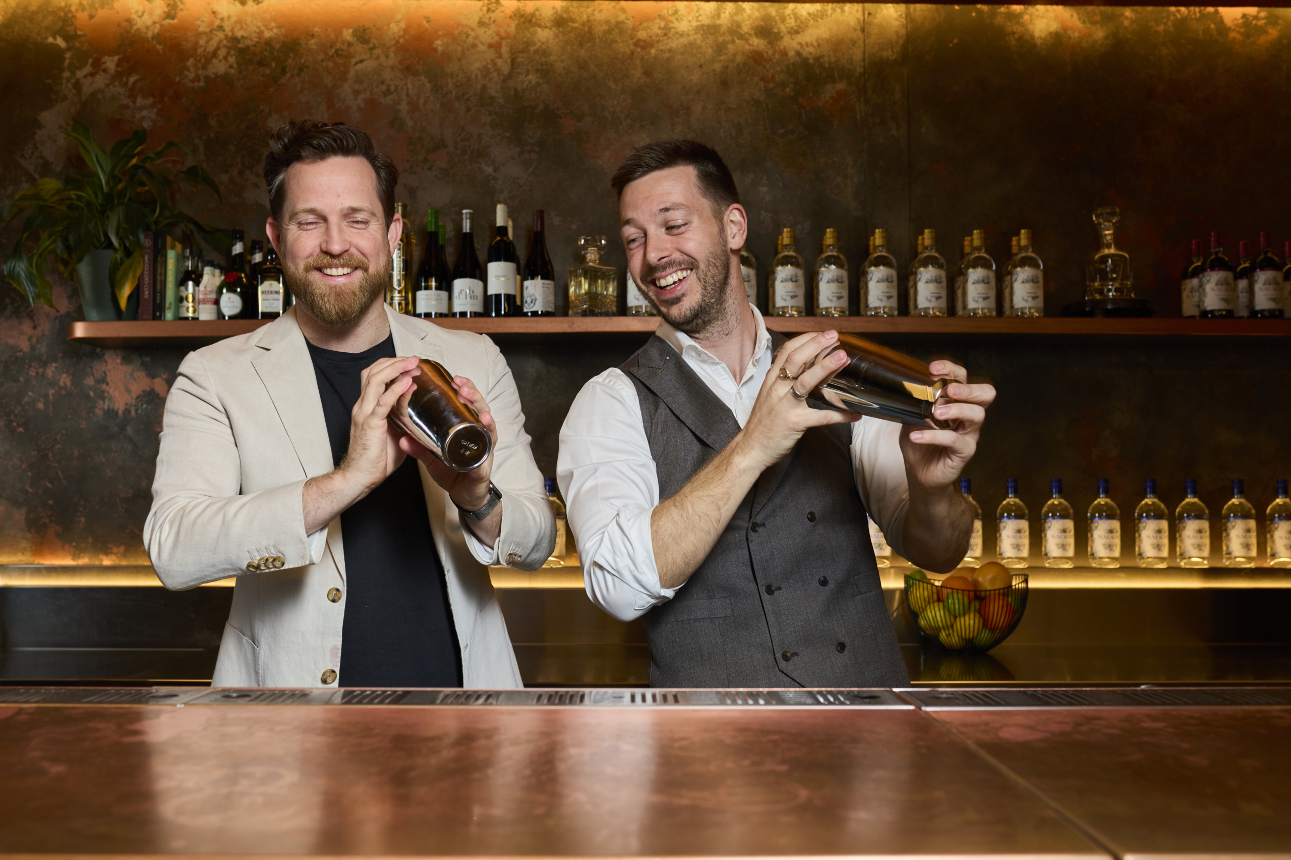 Two men smiling and shaking cocktail shakers behind a bar, with a backdrop of shelves filled with bottles, creating a lively and fun atmosphere at a cocktail-making session.