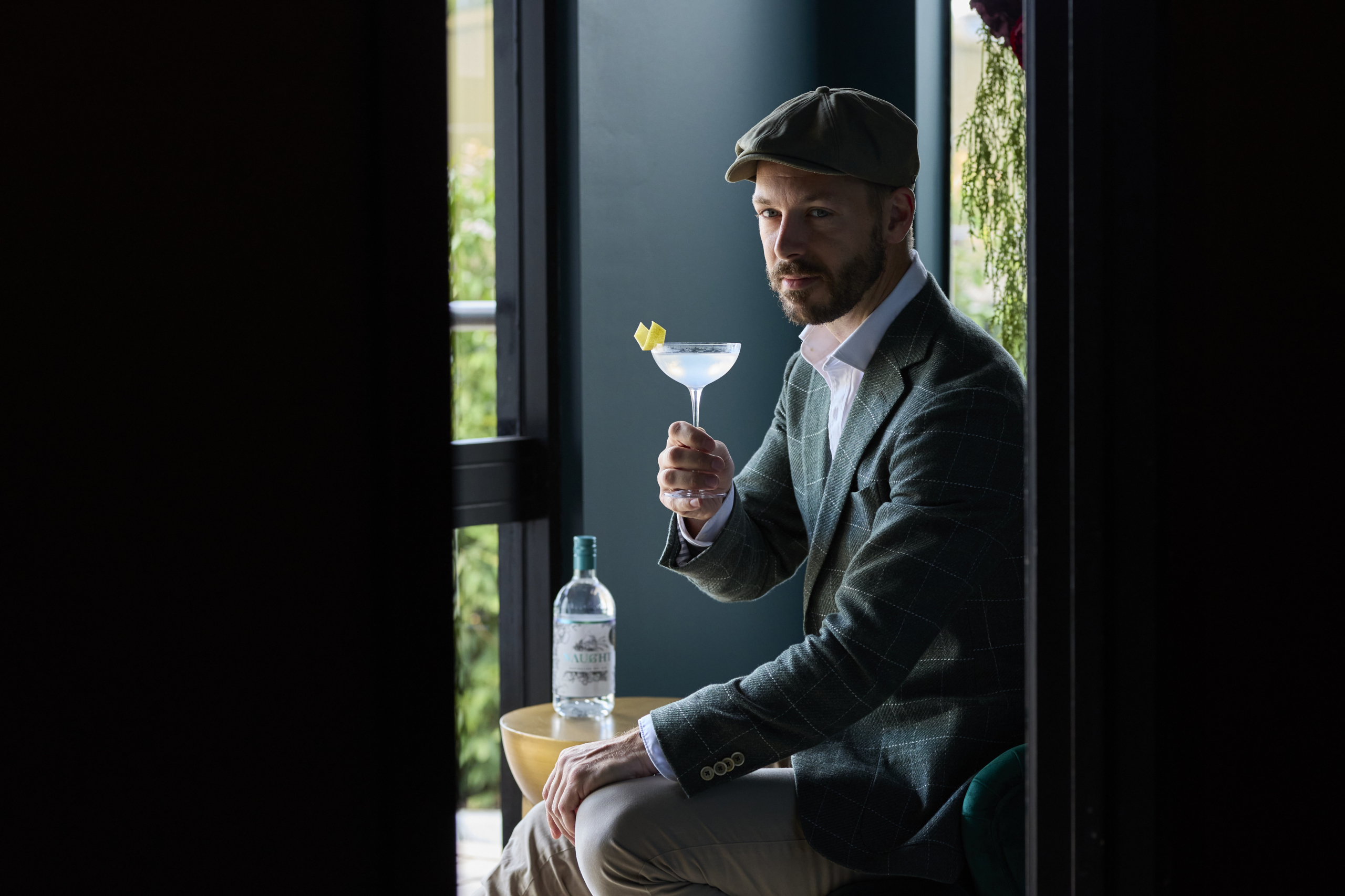 Man in a green checkered suit and flat cap sitting by a window, holding a cocktail glass with a lemon twist garnish, with a bottle of Naught gin on a table beside him, inviting you to join him for a moment of sophisticated relaxation.