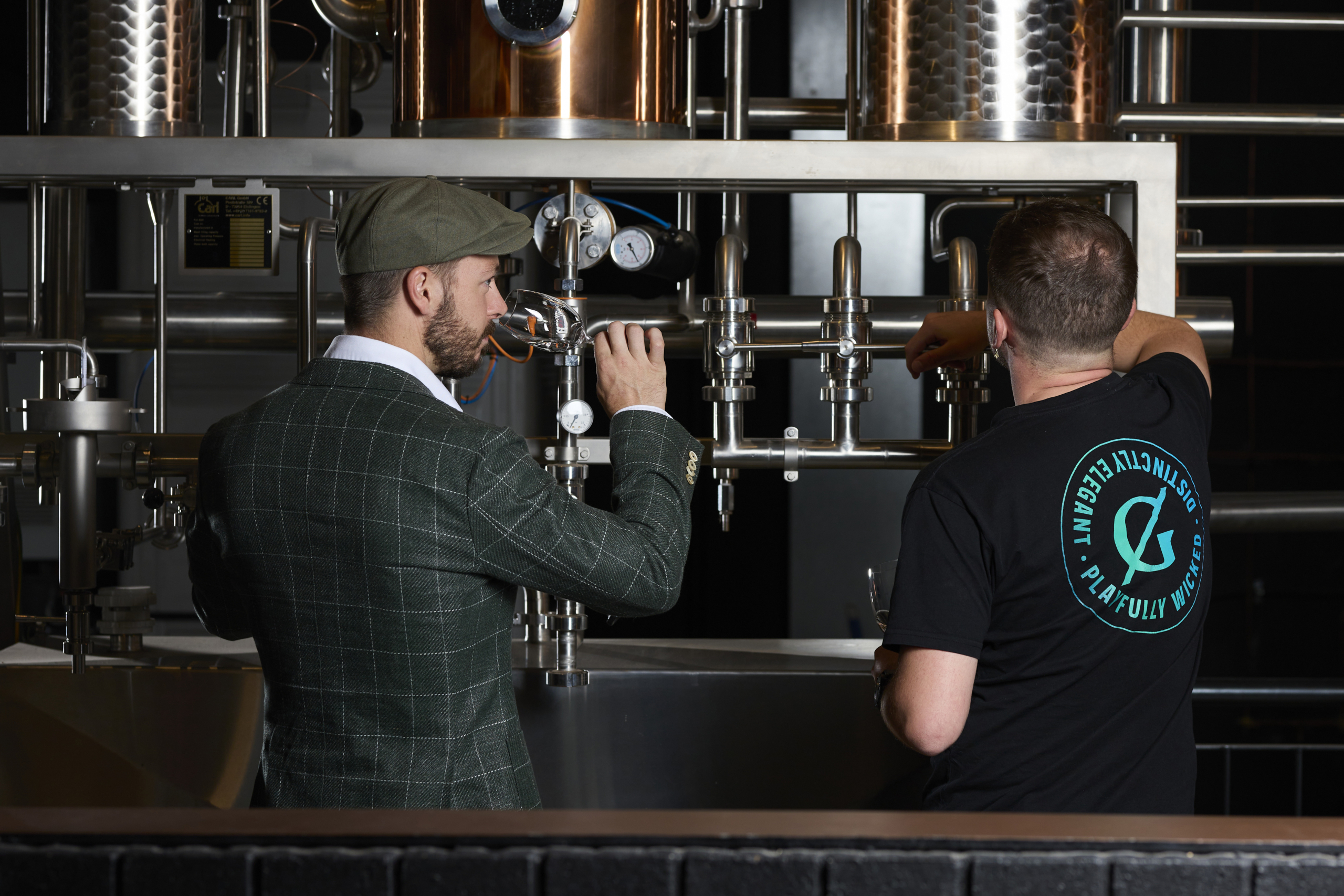 Two men working at a distillery, one in a checkered suit and flat cap tasting a spirit, while the other, wearing a branded Naught t-shirt, adjusts equipment, showcasing the craftsmanship and precision involved in gin production.