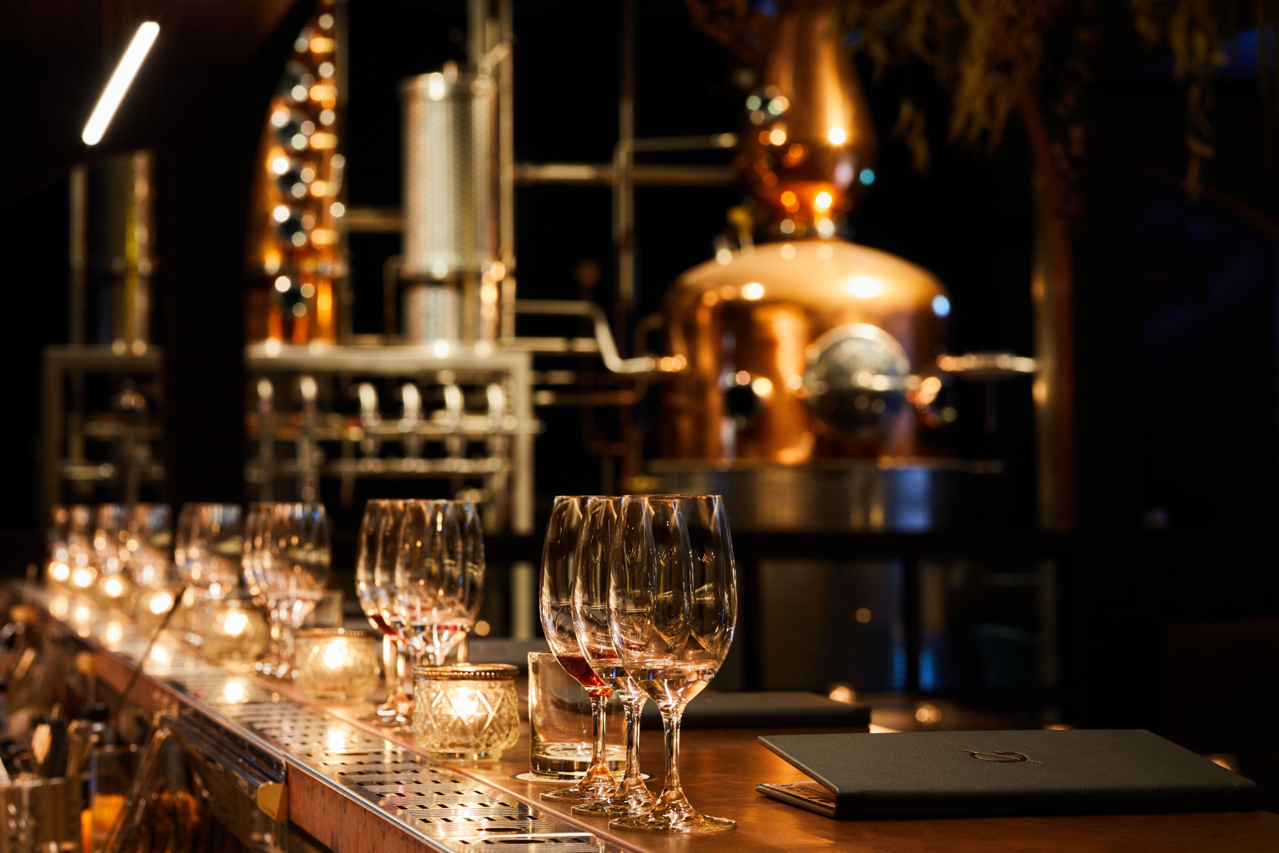 Elegant distillery interior with rows of wine glasses on a wooden bar counter, soft candlelight, and a backdrop of copper stills and intricate machinery, creating a warm, inviting ambiance for a tasting experience.